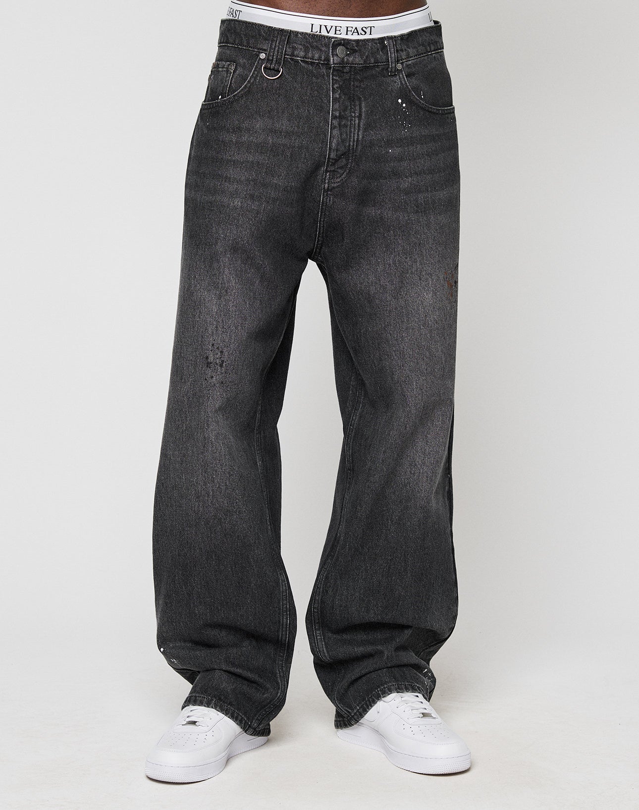 Wearing LFDY Baggy Denim by LIVE FAST DIE YOUNG, a person in baggy, faded black jeans with a wide straight leg and white sneakers stands by a light background; their underwear waistband reading LIVE FAST is visible above the jeans.