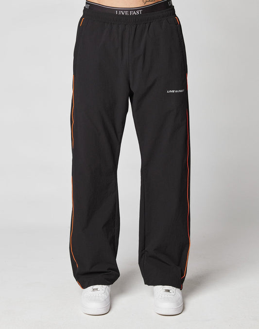 A person stands against a light background, wearing black LIVE FAST DIE YOUNG Live Fast Trackpants with thin orange side stripes, an elasticated drawstring waist, “LIVE FAST” text on the waistband and left thigh, and paired with white sneakers.