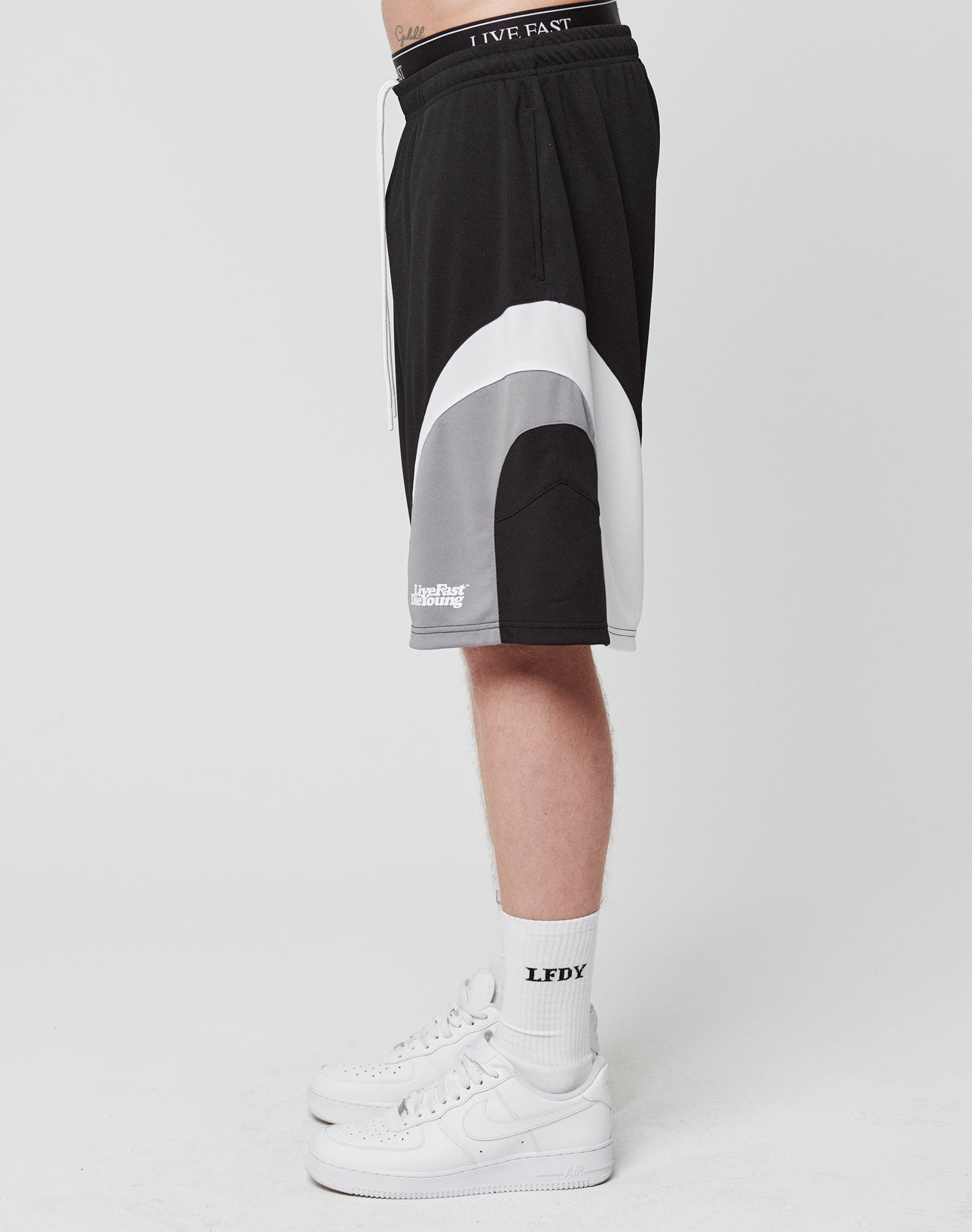 A person stands in profile against a plain white background, wearing LIVE FAST DIE YOUNG Basic Trackshorts with an elastic waistband, LFDY-branded white socks, and white Nike sneakers. Only the lower body is shown.