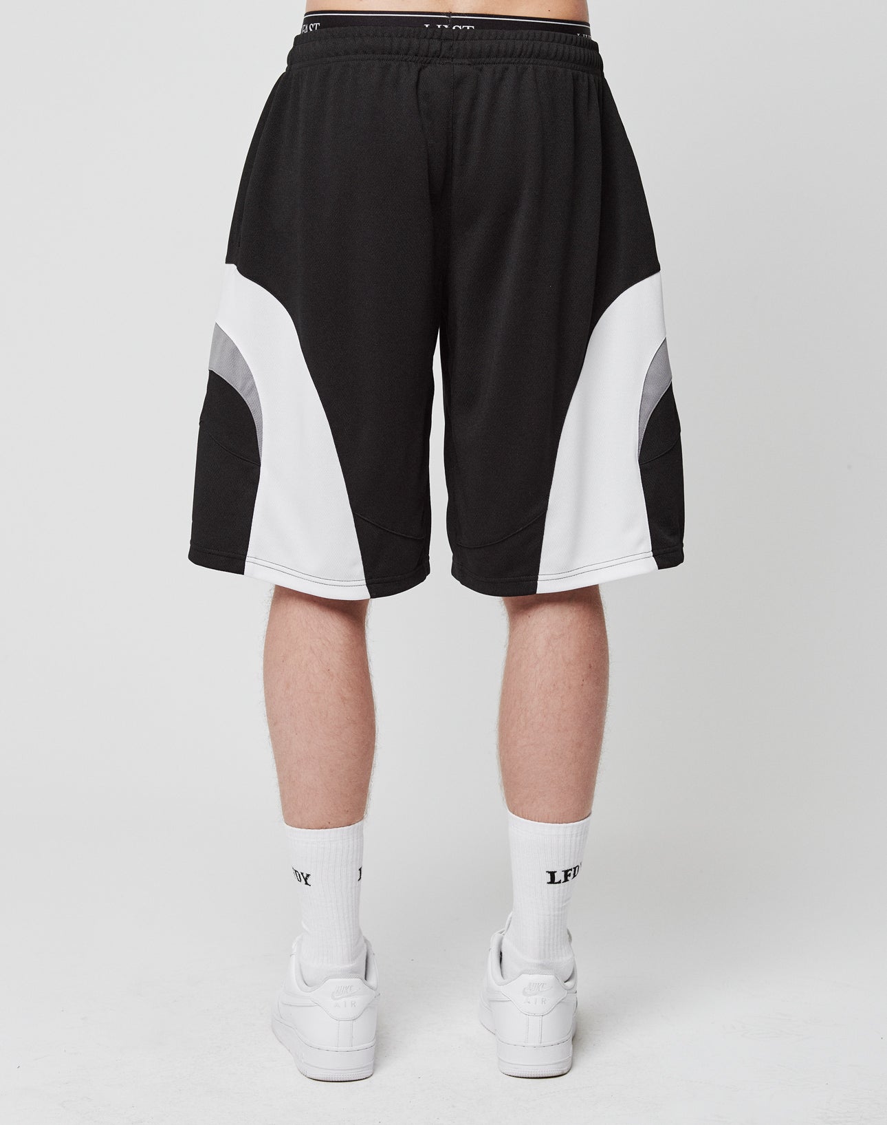 A person faces away from the camera wearing LIVE FAST DIE YOUNG Basic Trackshorts in a black and white logo print with elastic waistband, paired with white calf socks featuring black text and white sneakers, set against a plain background.