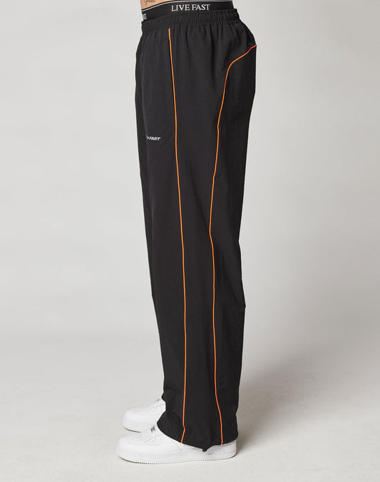 A person wears LIVE FAST DIE YOUNG Live Fast Trackpants in wide fit black with orange piping, reflective logo print, elastic waistband with LIVE FAST drawstring, and white sneakers. The side-view photo is set against a plain background.