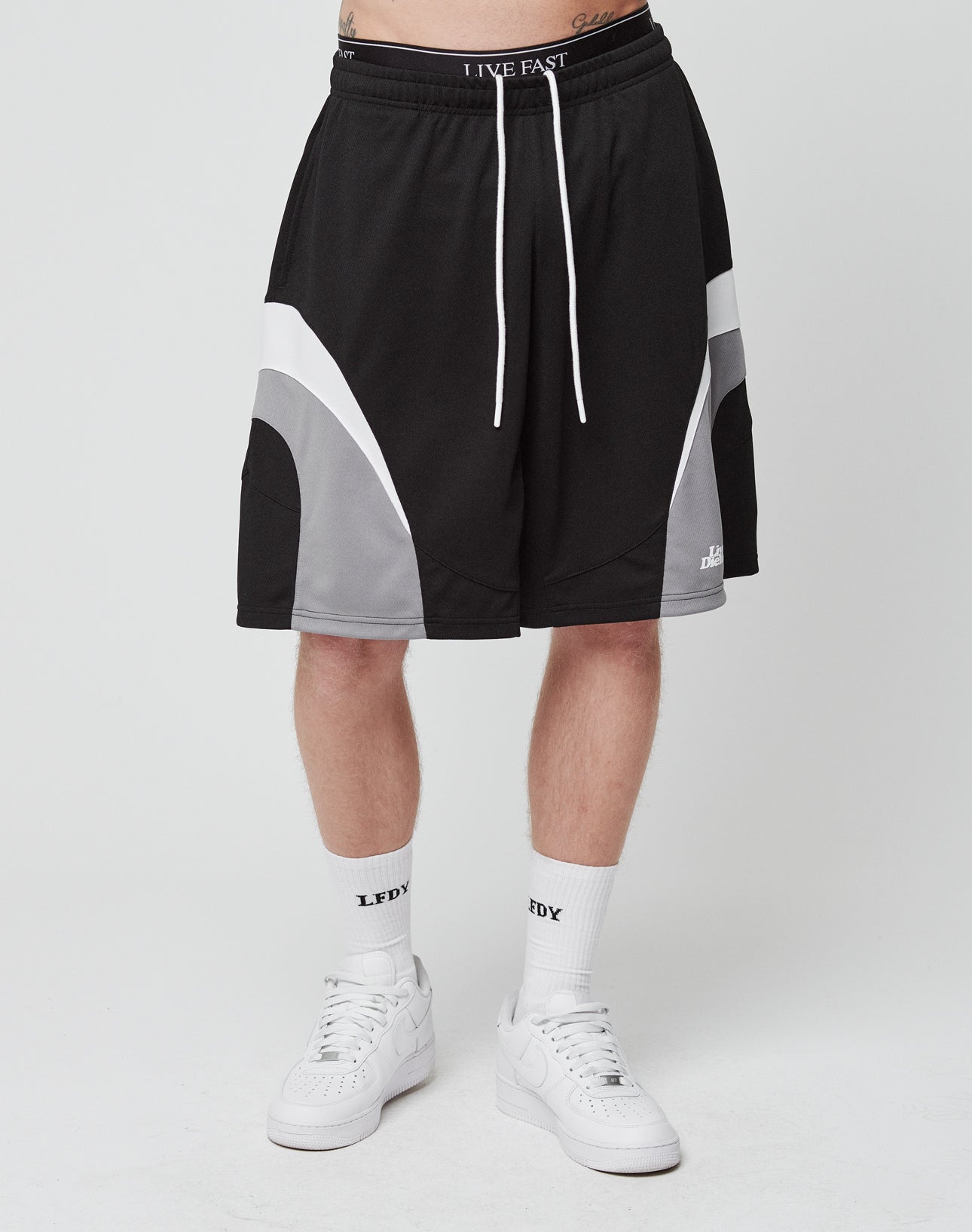 A person wears LIVE FAST DIE YOUNG Basic Trackshorts in black and gray with an elastic waistband and white drawstrings, LFDY logo-print white socks, and white sneakers. Only the lower half of the body is shown against a plain white background.