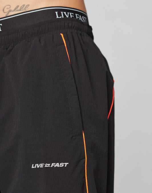 Close-up of a person wearing LIVE FAST DIE YOUNG Live Fast Trackpants: black wide-fit athletic shorts with LIVE FAST on the elastic waistband, orange piping on the pocket, and a reflective logo print. A waist tattoo is partly visible.
