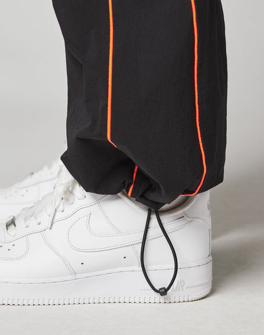 A close-up of a person wearing white Nike Air Force 1 sneakers and LIVE FAST DIE YOUNG Live Fast Trackpants in black with orange piping, elastic waistband with drawstring at the ankle, and a reflective logo print for added style.