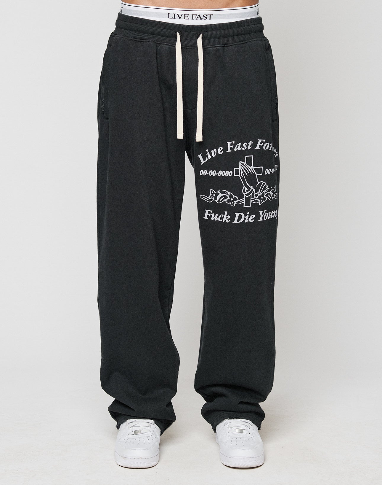 A person wears LFDY Praying Jogger sweatpants by LIVE FAST DIE YOUNG, featuring an elastic waistband with white drawstrings, white sneakers, and oversized fit with Live Fast Forever text and graphic details.