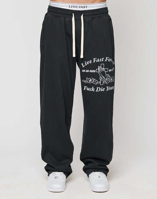 A person wears LFDY Praying Jogger sweatpants by LIVE FAST DIE YOUNG, featuring an elastic waistband with white drawstrings, white sneakers, and oversized fit with Live Fast Forever text and graphic details.