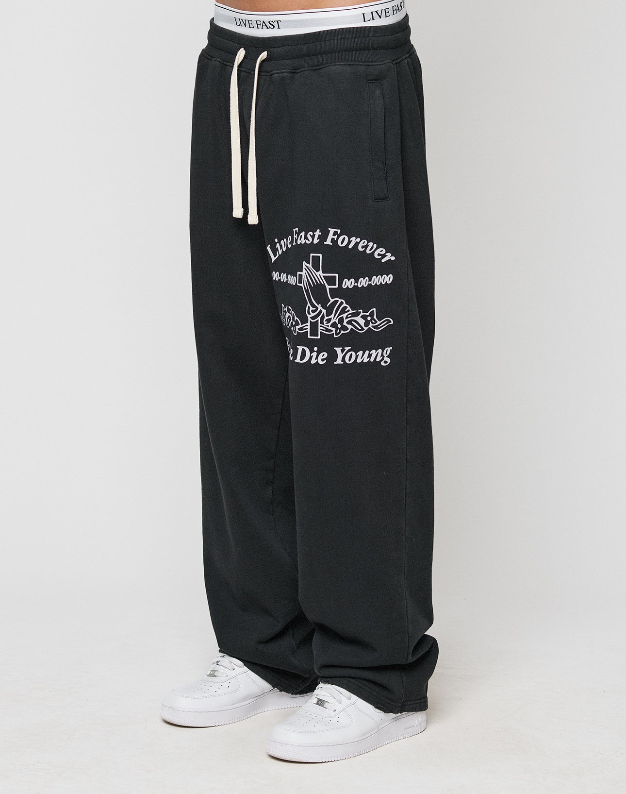 A person wearing LIVE FAST DIE YOUNGs LFDY Praying Jogger—black baggy sweatpants with white graphics, an oversized fit, and elastic waistband—paired with white sneakers. Only the lower body is visible.