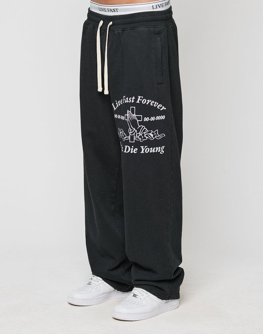 A person wearing LIVE FAST DIE YOUNGs LFDY Praying Jogger—black baggy sweatpants with white graphics, an oversized fit, and elastic waistband—paired with white sneakers. Only the lower body is visible. | model
