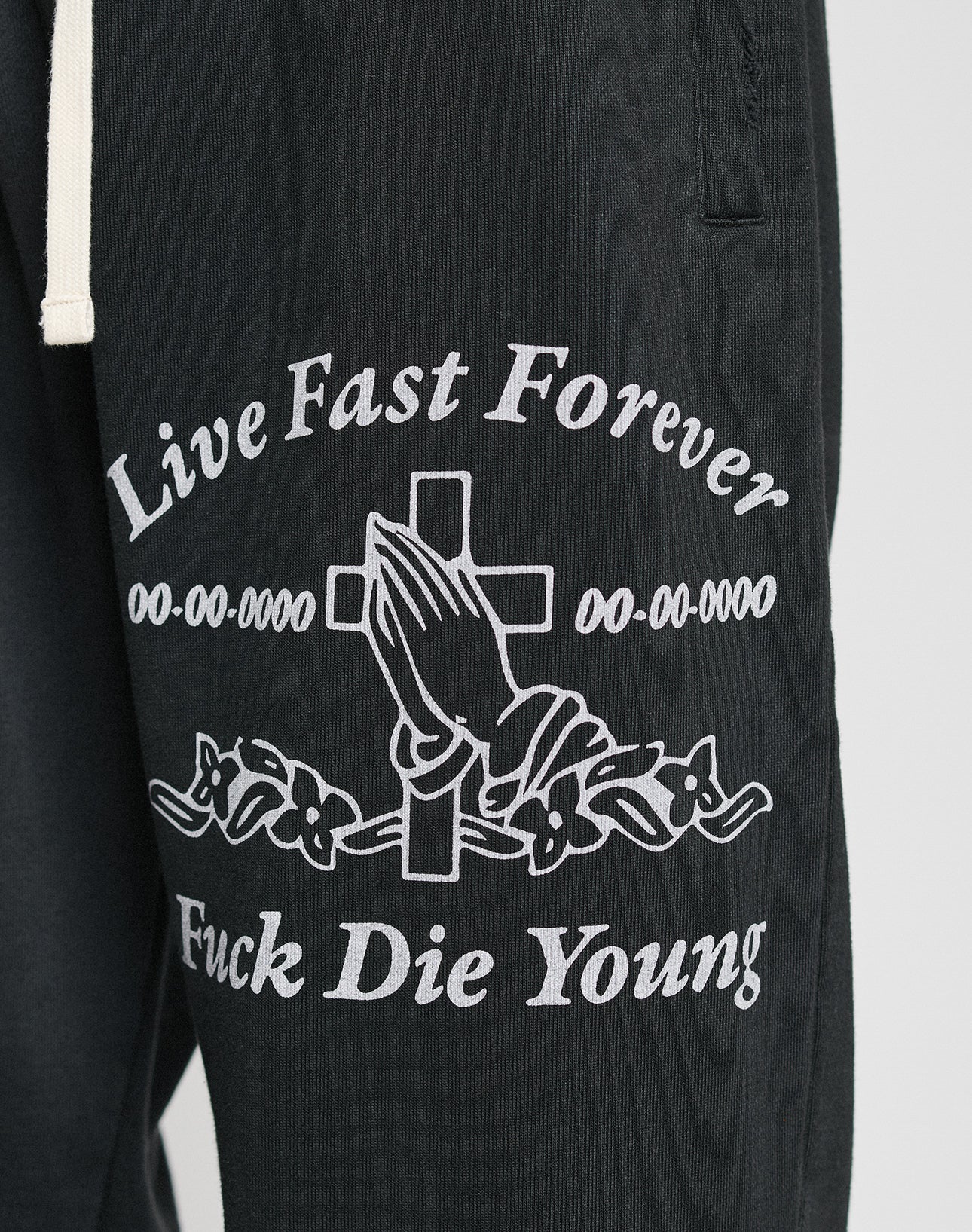 The LFDY Praying Jogger by LIVE FAST DIE YOUNG features black sweatpants with an elastic waistband, white praying hands graphic in front of a cross, “Live Fast Forever” & “Fuck Die Young” text, floral accents, and a relaxed oversize fit.