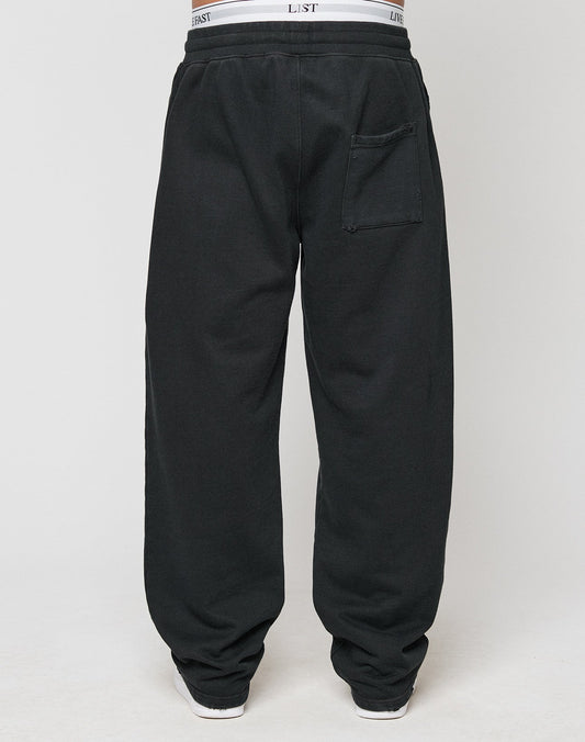 A person is seen from the back wearing LIVE FAST DIE YOUNG’s LFDY Praying Jogger—loose-fitting black sweatpants with an oversize fit, a visible back pocket, and white underwear waistband peeking over the elastic waist.