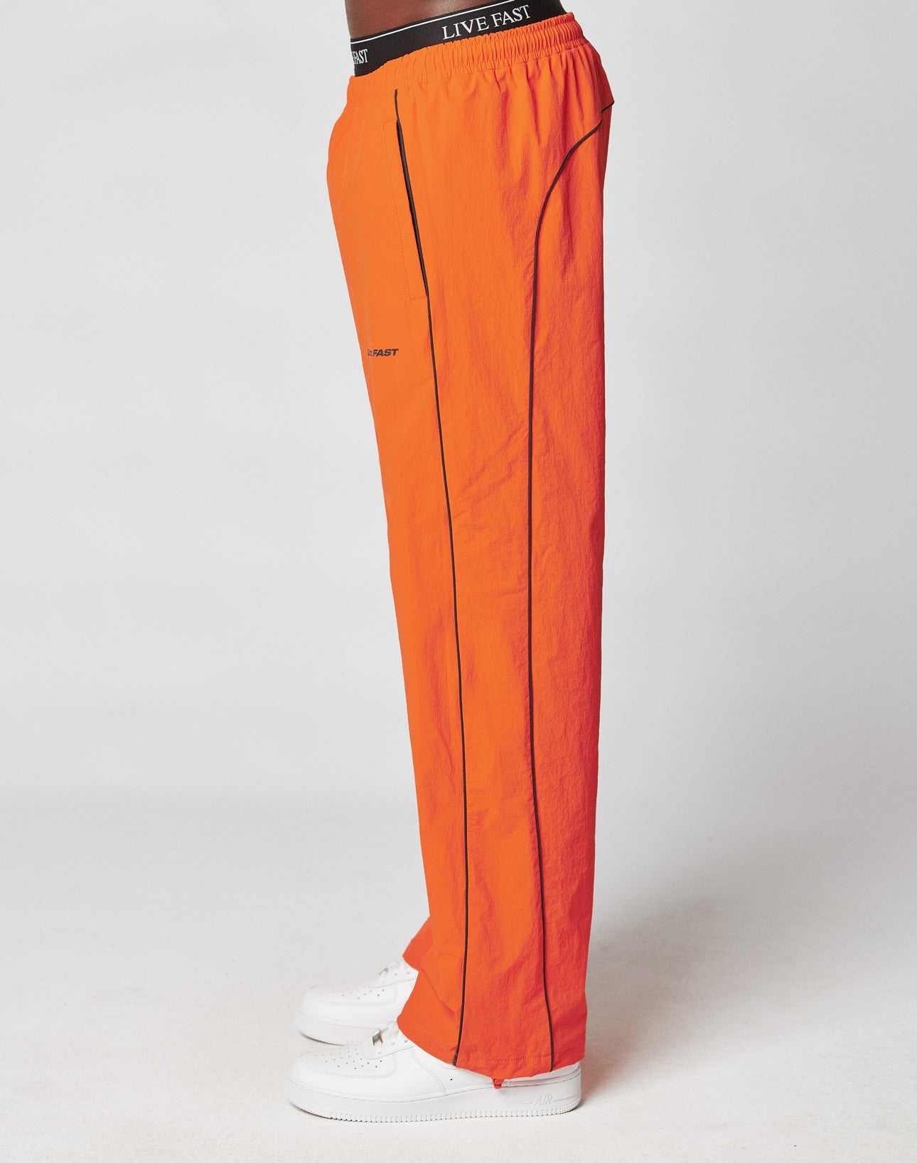 A person models LIVE FAST DIE YOUNG’s bright orange Live Fast Trackpants with a Breiter Elastik-Bund, black piping, and LIVE FAST on the waistband, paired with white sneakers against a white background.