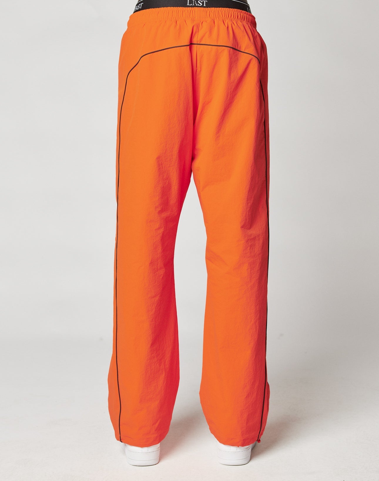 A person is seen from the back wearing LIVE FAST DIE YOUNG’s bright orange Live Fast Trackpants with black piping, a wide elastic waistband, and a reflective logo print, set against a plain light background.