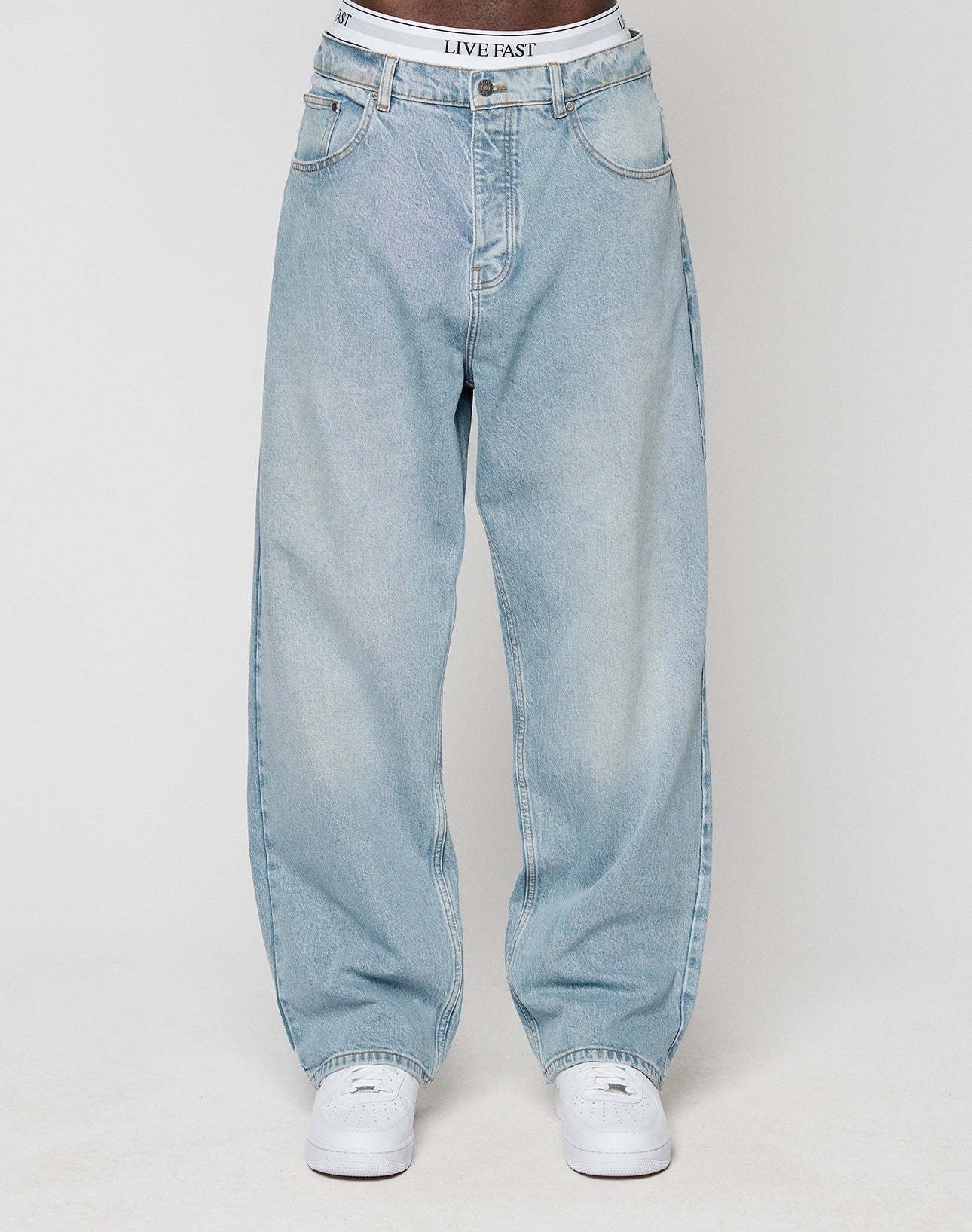 A person wearing light blue LFDY Baggy Denim by LIVE FAST DIE YOUNG, white sneakers, and a visible white waistband with LIVE FAST text, stands against a plain light background.