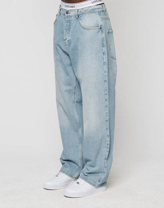 Someone wears loose-fitting, light blue LFDY Baggy Denim jeans by LIVE FAST DIE YOUNG with a gerades Bein and baggy fit, white sneakers, and a visible “LIVE FAST” waistband. The background is plain and light-colored. | model
