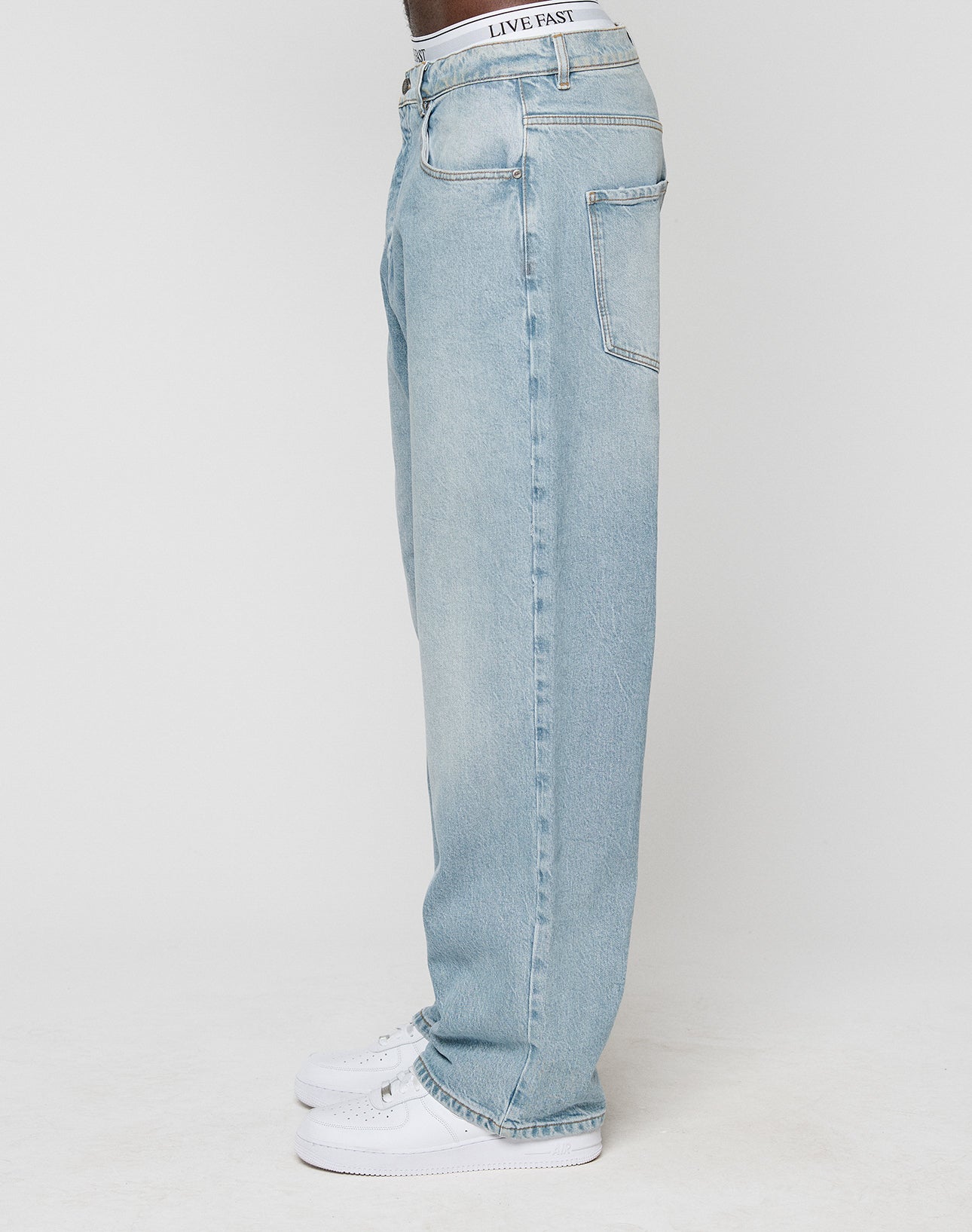 A person wears LFDY Baggy Denim by LIVE FAST DIE YOUNG in light blue, 100% Baumwolle, with a baggy fit and straight leg. They stand sideways with white sneakers, only their lower body visible against a plain white background.