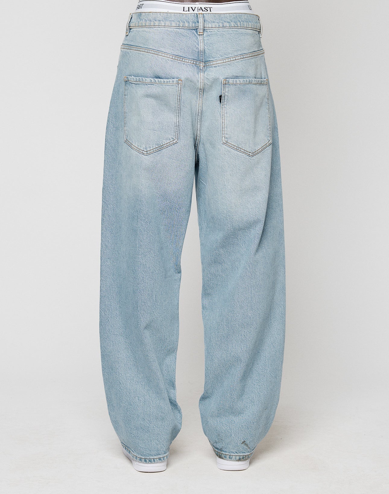 The LFDY Baggy Denim by LIVE FAST DIE YOUNG features a light blue baggy fit, two large rear pockets, a straight leg design, and is made from 100% cotton. A white waistband accents the top with white shoes visible below.