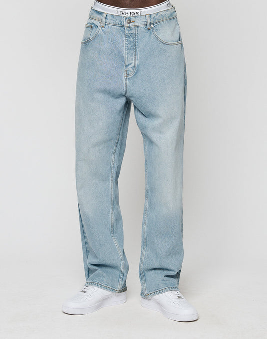 Wearing LFDY Baggy Denim by LIVE FAST DIE YOUNG in light blue, with LIVE FAST on the white waistband, gerades Bein style, 100% Baumwolle—paired with white sneakers against a plain light background.
