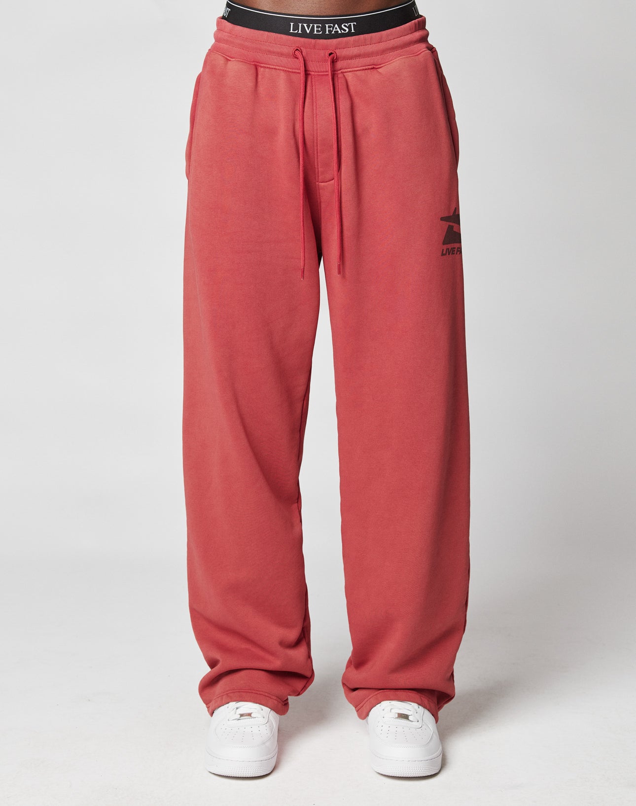 A person wears the Live Fast Sweatsuit Jogger by LIVE FAST DIE YOUNG in a loose, Oversize Fit with an Elastik-Bund waistband showing “LIVE FAST,” a small logo on the left thigh, and white sneakers, posed against a plain background.