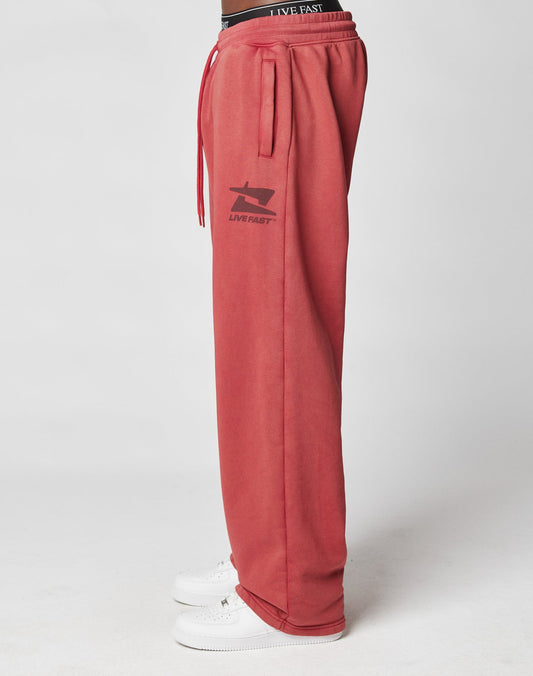 A person wearing LIVE FAST DIE YOUNG’s Live Fast Sweatsuit Jogger—loose-fitting, vintage wash red with a thigh logo, black waistband, and white sneakers—is shown from the side against a plain white background.