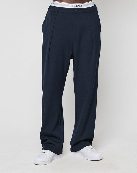 A person wears LIVE FAST DIE YOUNG Wide Casual Pants in dark navy with a visible white waistband featuring LIVE FAST text, paired with white athletic shoes and styled in a straight-leg silhouette against a plain light background.