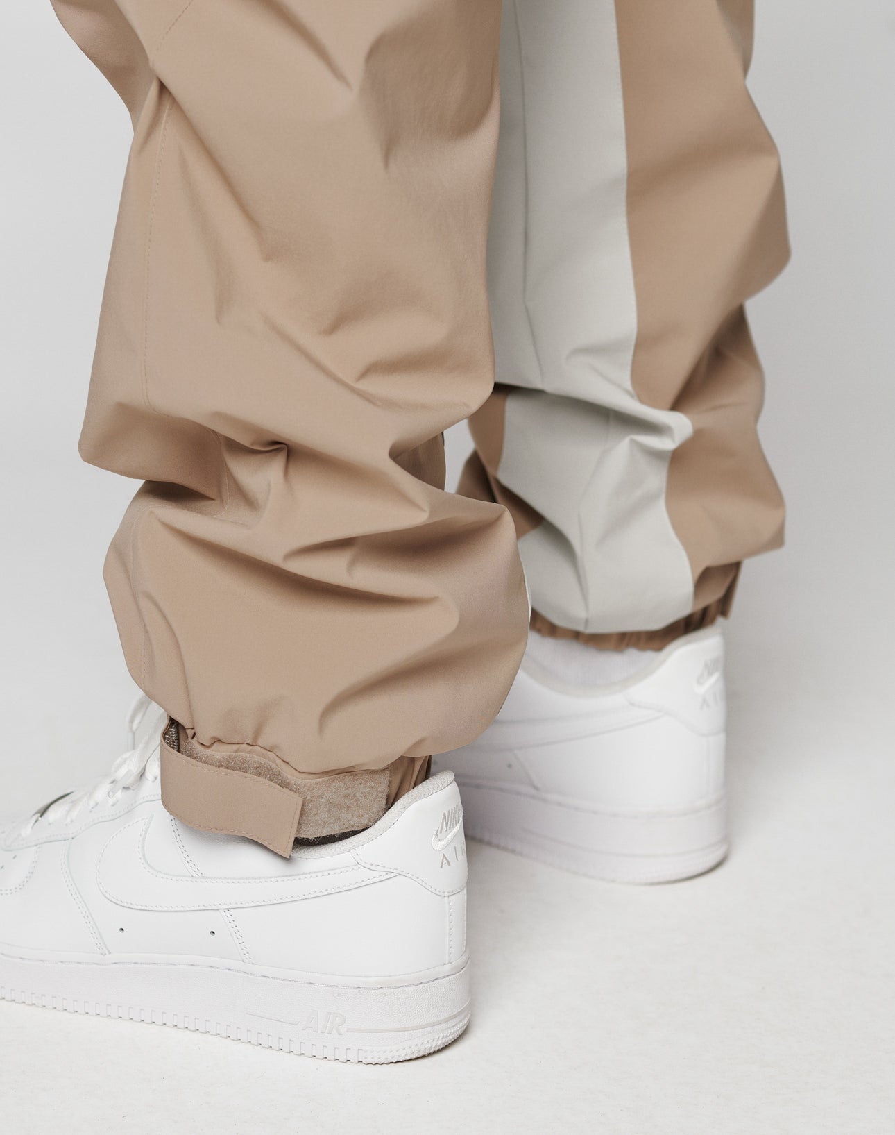 Close-up of a person wearing LIVE FAST DIE YOUNG Basic Shell Pants in beige and light gray with elastic waistband and drawcord, paired with white Nike Air Force 1 sneakers, standing on a light surface.