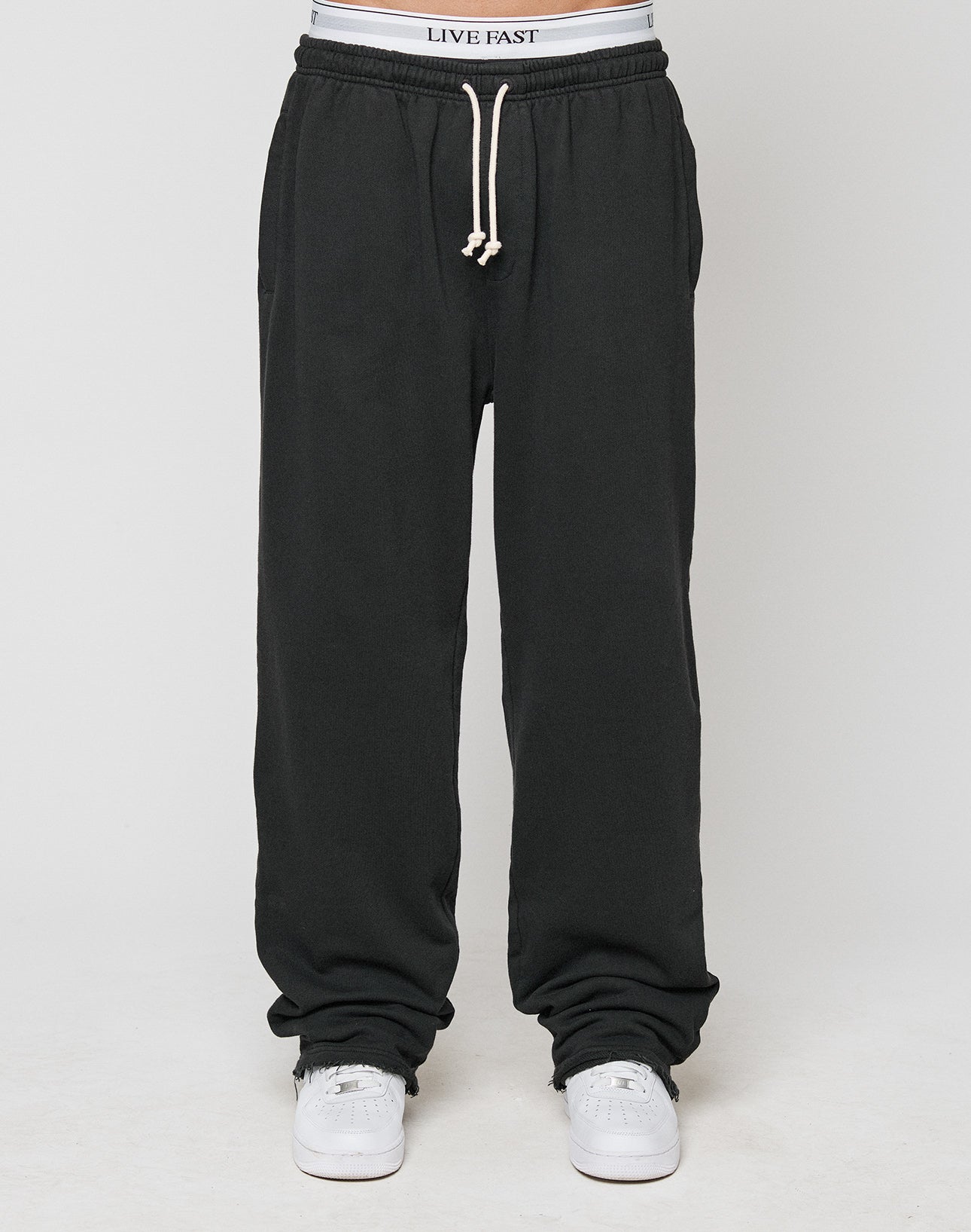A person wears LIVE FAST DIE YOUNGs LFDY Lightweight Jogger—loose black sweatpants with an elastic waistband reading LIVE FAST, made of soft French Terry—paired with white sneakers and a white drawstring, against a plain white background.