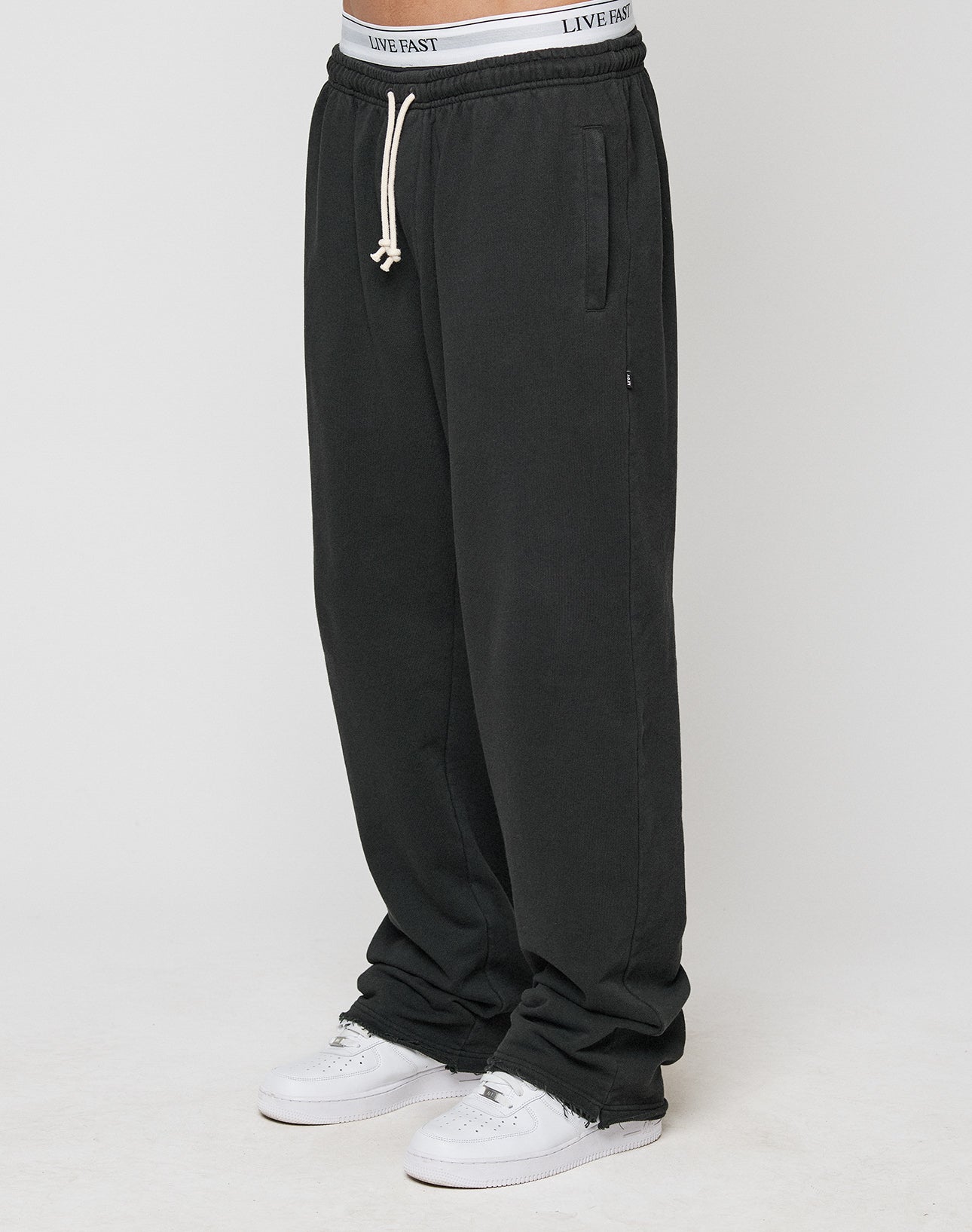 A person in LIVE FAST DIE YOUNG LFDY Lightweight Joggers with elastic waistband and drawstring, paired with white sneakers and a visible white LIVE FAST waistband, stands against a simple light background. | model