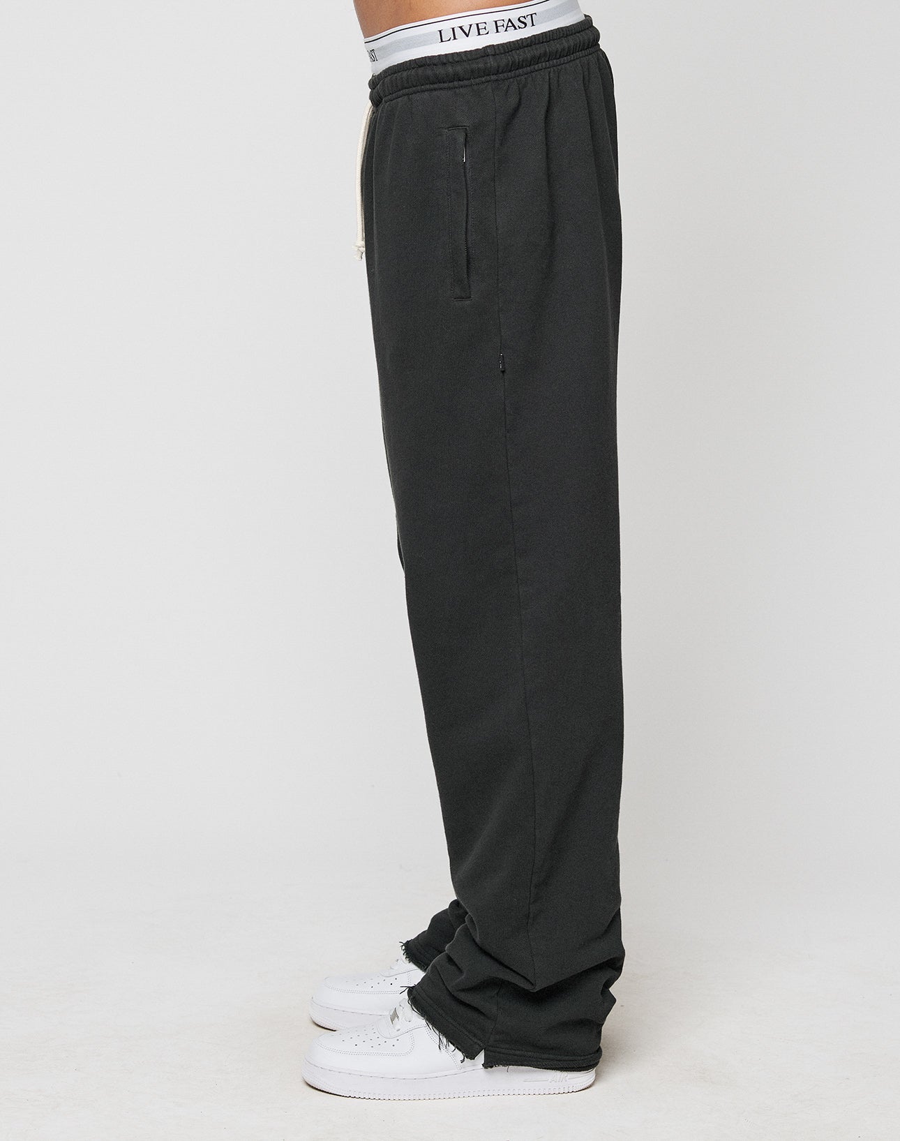 A person stands in profile wearing oversize fit black LFDY Lightweight Jogger pants by LIVE FAST DIE YOUNG, featuring soft French terry fabric and a gray waistband with LIVE FAST text, paired with white sneakers against a plain white background.