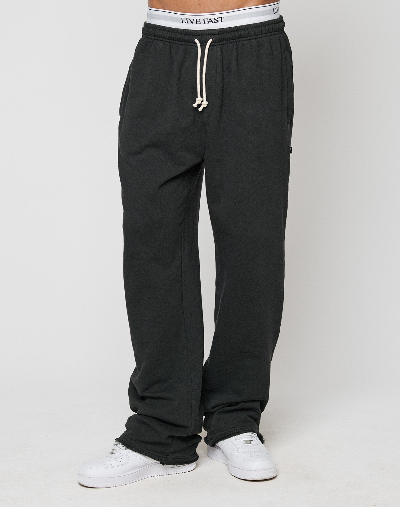 A person wears LFDY Lightweight Jogger by LIVE FAST DIE YOUNG in oversize fit black with white drawstrings, paired with white sneakers and a visible LIVE FAST waistband, standing against a plain white background.