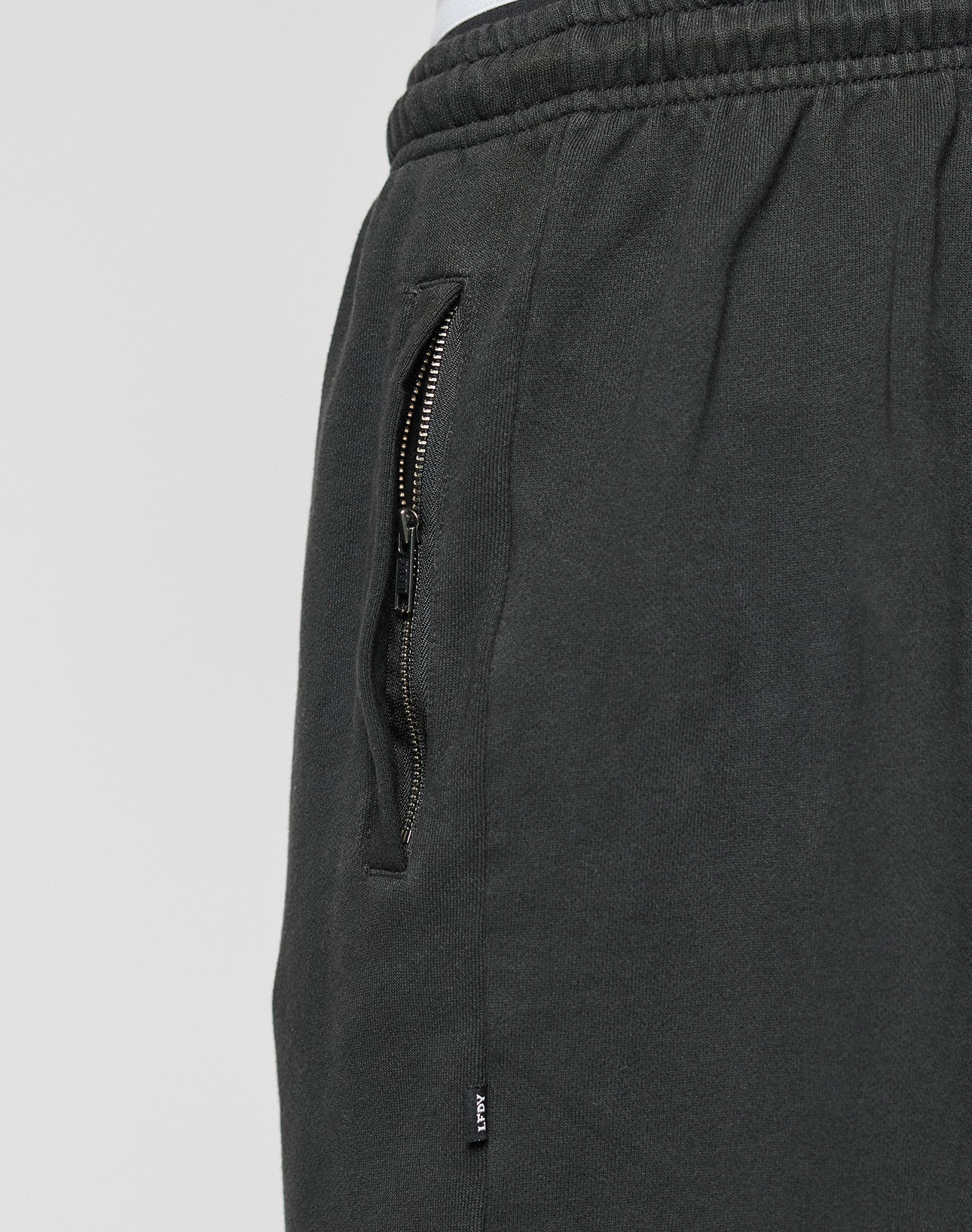 Close-up of the LFDY Lightweight Jogger by LIVE FAST DIE YOUNG, showing its dark gray French Terry fabric, elastikbund waistband, zippered side pocket, and a small tag near the pocket. Oversize fit is highlighted.