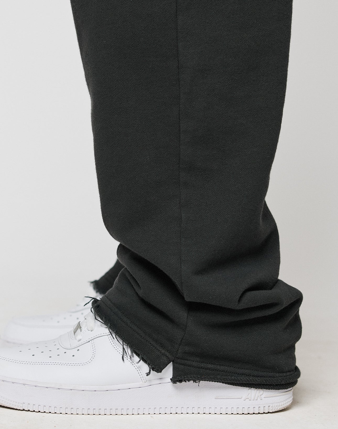 A close-up side view of a person wearing the LIVE FAST DIE YOUNG LFDY Lightweight Jogger in black, featuring frayed hems and an elastic waistband in an oversize fit over white sneakers, standing on a light surface.