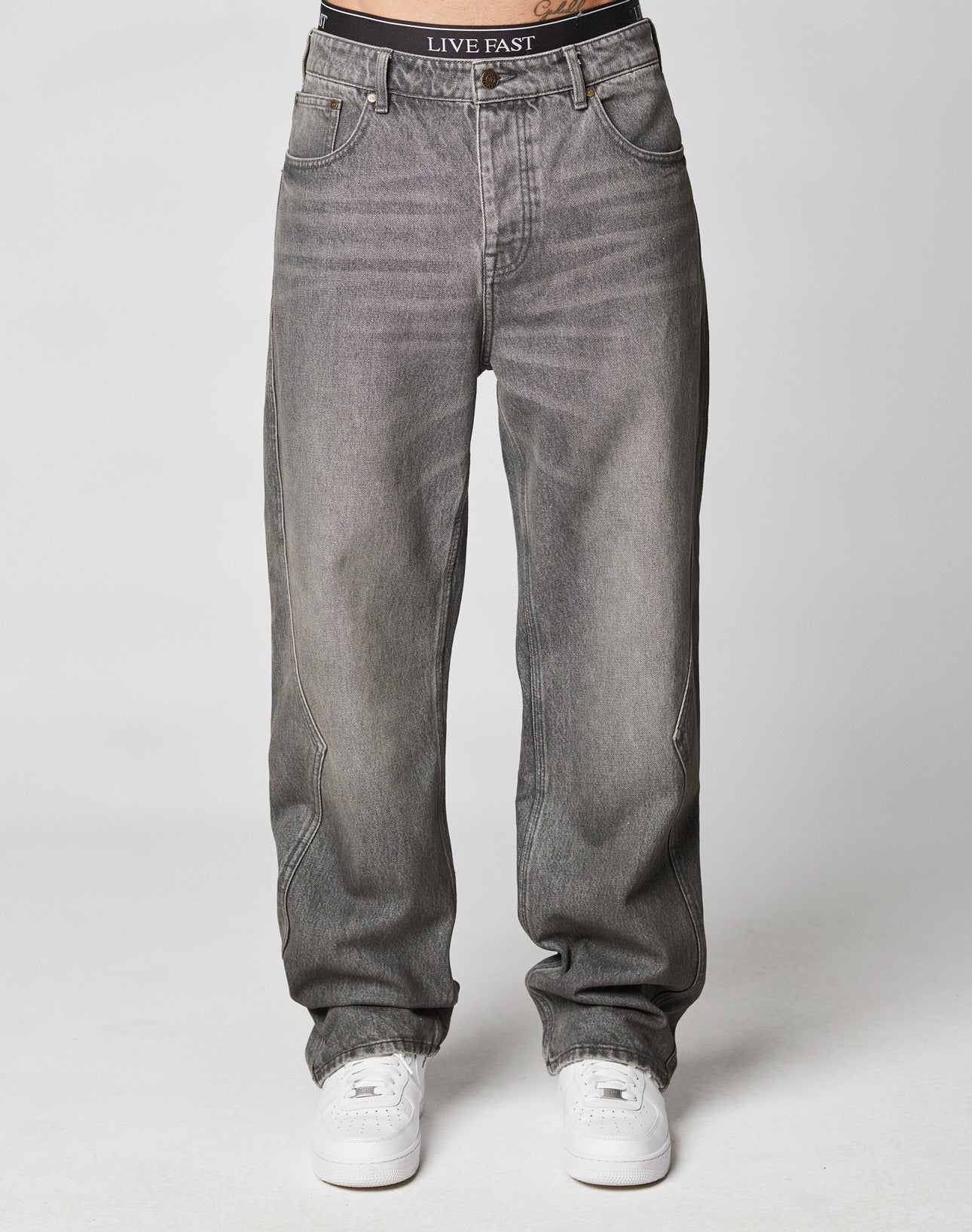 A person wears gray Rhomb Wide Denim jeans by LIVE FAST DIE YOUNG, featuring a baggy fit and visible waistband label, with white sneakers. The jeans have a wide straight leg, and the torso isnt shown, set against a plain light background.