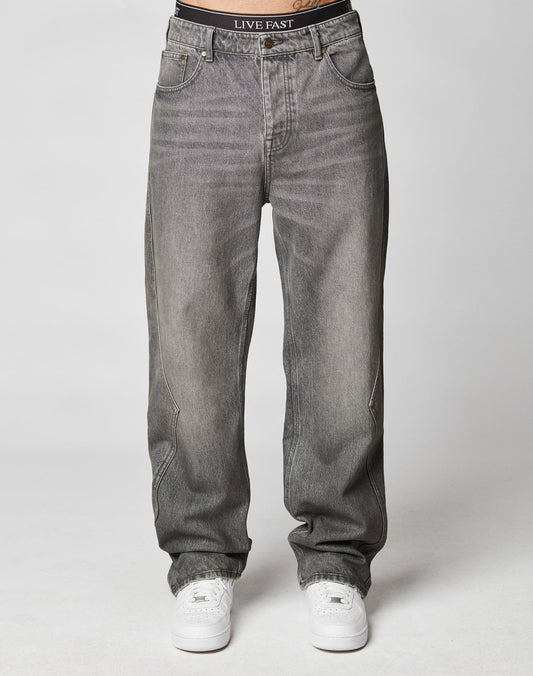A person wears gray Rhomb Wide Denim jeans by LIVE FAST DIE YOUNG, featuring a baggy fit and visible waistband label, with white sneakers. The jeans have a wide straight leg, and the torso isnt shown, set against a plain light background.