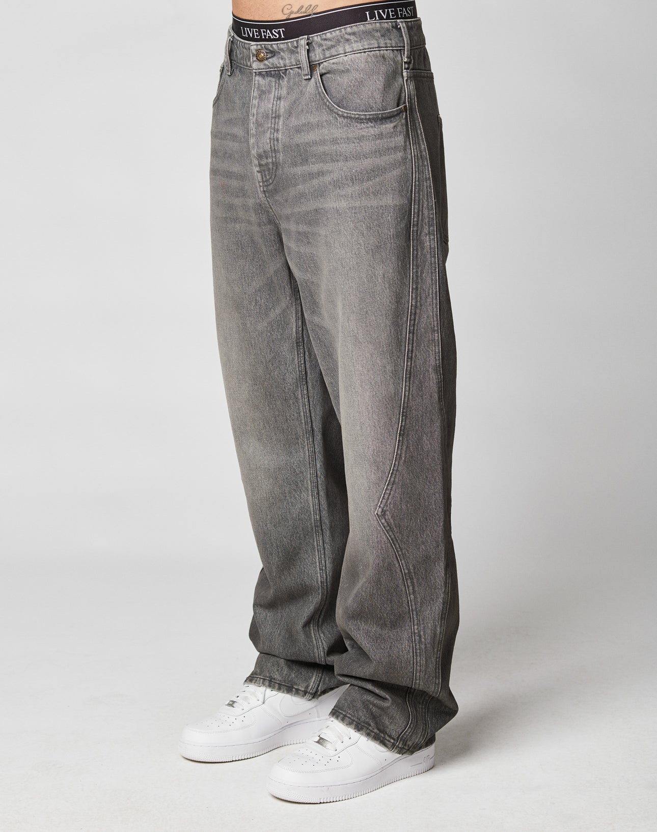A person in baggy Rhomb Wide Denim by LIVE FAST DIE YOUNG, loose gray jeans with wide straight legs, and white sneakers stands against a plain background. The waistband of black LIVE FAST underwear shows above the 100% cotton jeans.