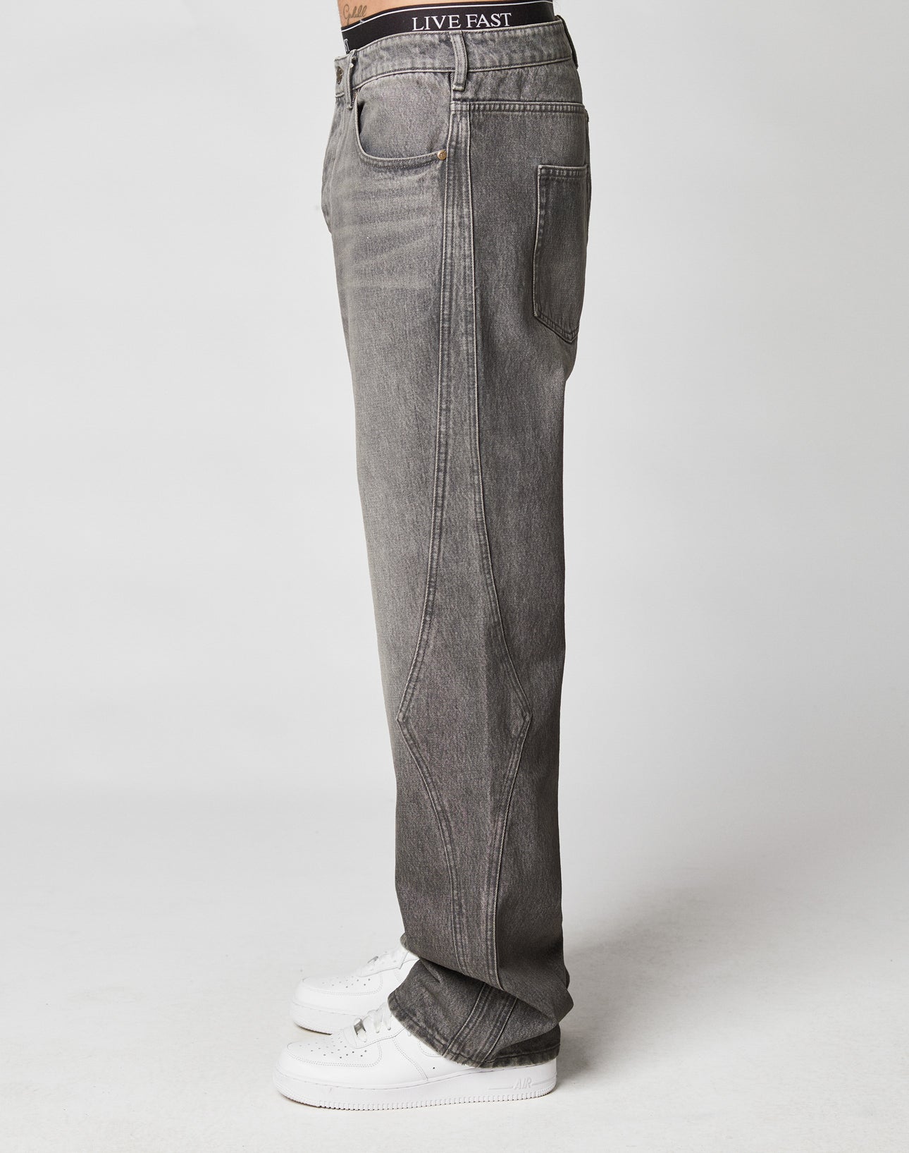 A person in Rhomb Wide Denim by LIVE FAST DIE YOUNG, gray baggy jeans with a wide straight leg made of 100% cotton, white sneakers, and a black LIVE FAST waistband stands sideways against a white background.