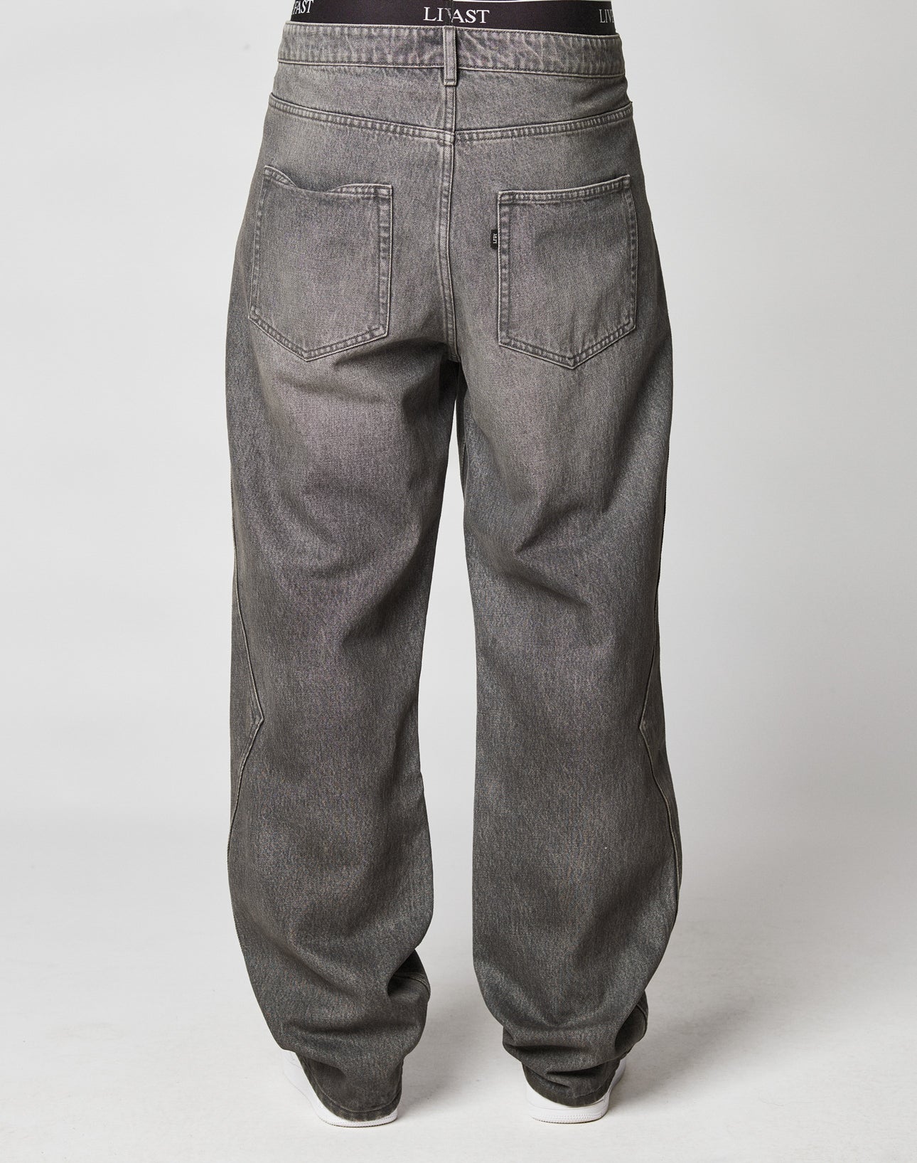 Someone wears Rhomb Wide Denim by LIVE FAST DIE YOUNG—baggy faded gray jeans (100% cotton, wide straight leg, two back pockets). A black waistband with a visible LIAST logo peeks above the jeans against a plain light background.