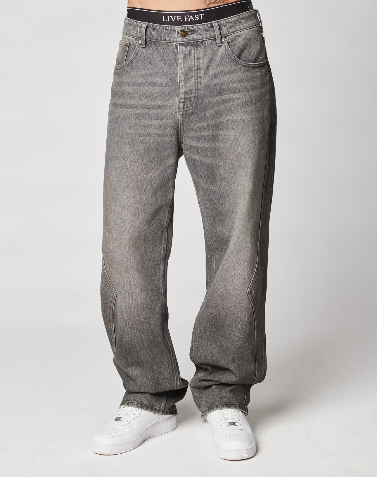 Someone wears the Rhomb Wide Denim by LIVE FAST DIE YOUNG, featuring a gray baggy fit with weitem geradem Bein, white sneakers, and black underwear with LIVE FAST on the waistband, standing against a plain light background.