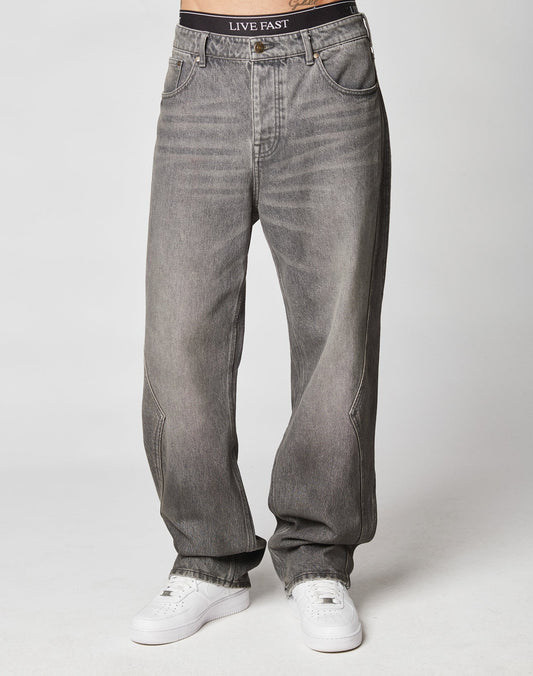 Someone wears the Rhomb Wide Denim by LIVE FAST DIE YOUNG, featuring a gray baggy fit with weitem geradem Bein, white sneakers, and black underwear with LIVE FAST on the waistband, standing against a plain light background.