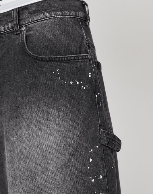 Close-up of LIVE FAST DIE YOUNG Carpenter Jorts in black, baggy fit, 100% cotton with white paint-splatter details and a side utility pocket, shown from waist to mid-thigh against a plain background.