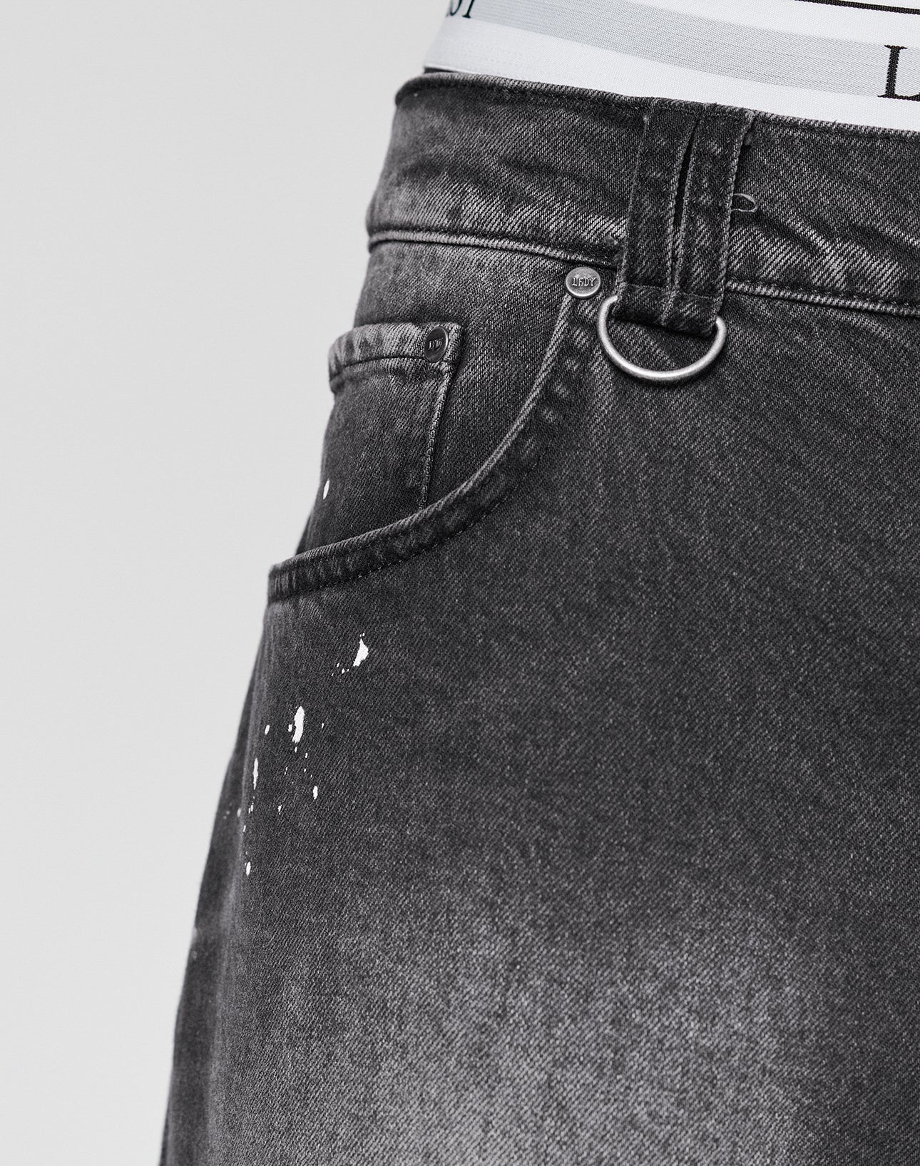 Close-up of a person wearing LIVE FAST DIE YOUNG Carpenter Jorts in dark gray, Baggy Fit, with paint splatter near the front pocket, a silver keyring on the belt loop, and white underwear waistband visible above the shorts.