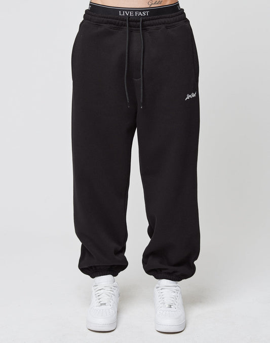 A person wearing black Loose Jogger sweatpants by LIVE FAST DIE YOUNG, featuring an elastic waistband with LIVE FAST branding, elastic cuffs, small white script on the left thigh, and white sneakers, stands against a plain light background.