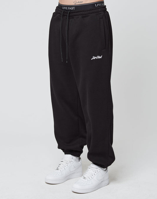 A person wears black Loose Jogger sweatpants by LIVE FAST DIE YOUNG, featuring Live Fast embroidery on the left thigh, elastic cuffs, a black waistband with Live Fast, and soft Baumwolle fabric. White Nike sneakers complete the look against a plain white background. | model