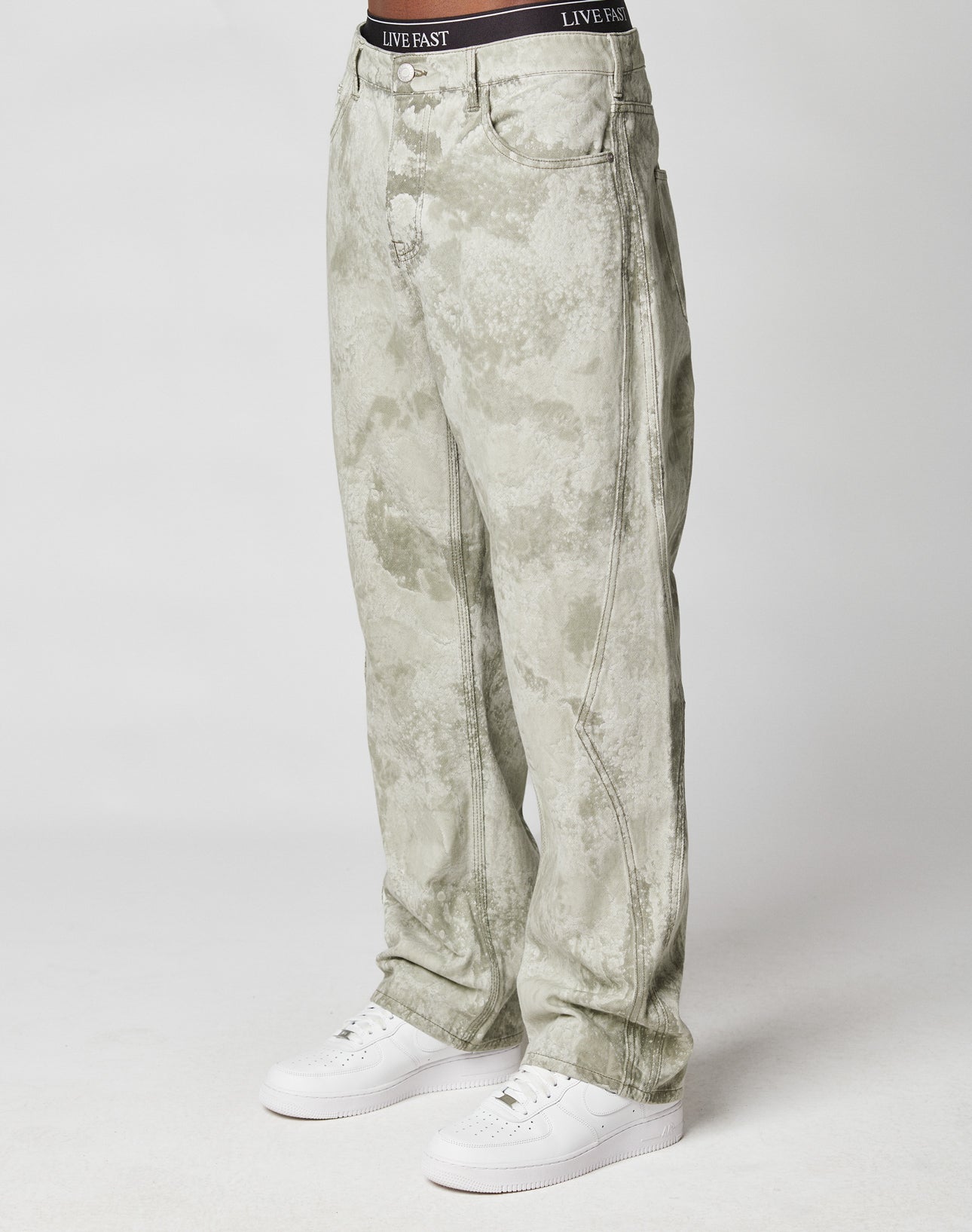 A person models the Rhomb Wide Denim by LIVE FAST DIE YOUNG—loose, light gray acid-wash jeans with diamond-shaped panels, visible seams, a black branded waistband, and a baggy fit—against a white background. Made from 100% Baumwolle.