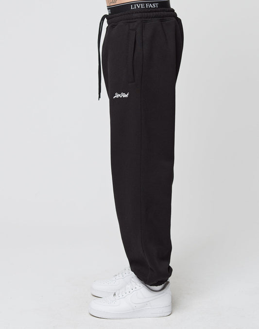 A person stands sideways in black LIVE FAST DIE YOUNG Loose Jogger sweatpants with Live Fast waistband, white thigh embroidery, elastic ankle cuffs, and white Nike sneakers against a plain white background.