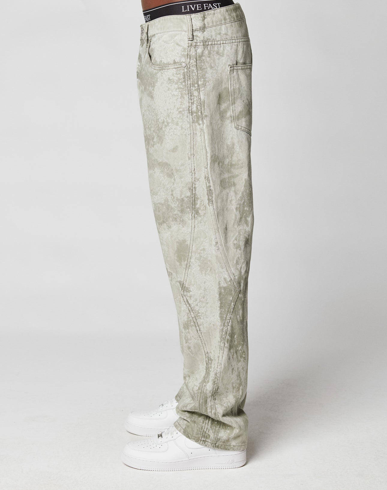 A person in profile wears the Rhomb Wide Denim by LIVE FAST DIE YOUNG—acid-wash, light gray baggy jeans with diamond-shaped panels—paired with white sneakers and black “LIVE FAST” waistband underwear, set against a plain white background.