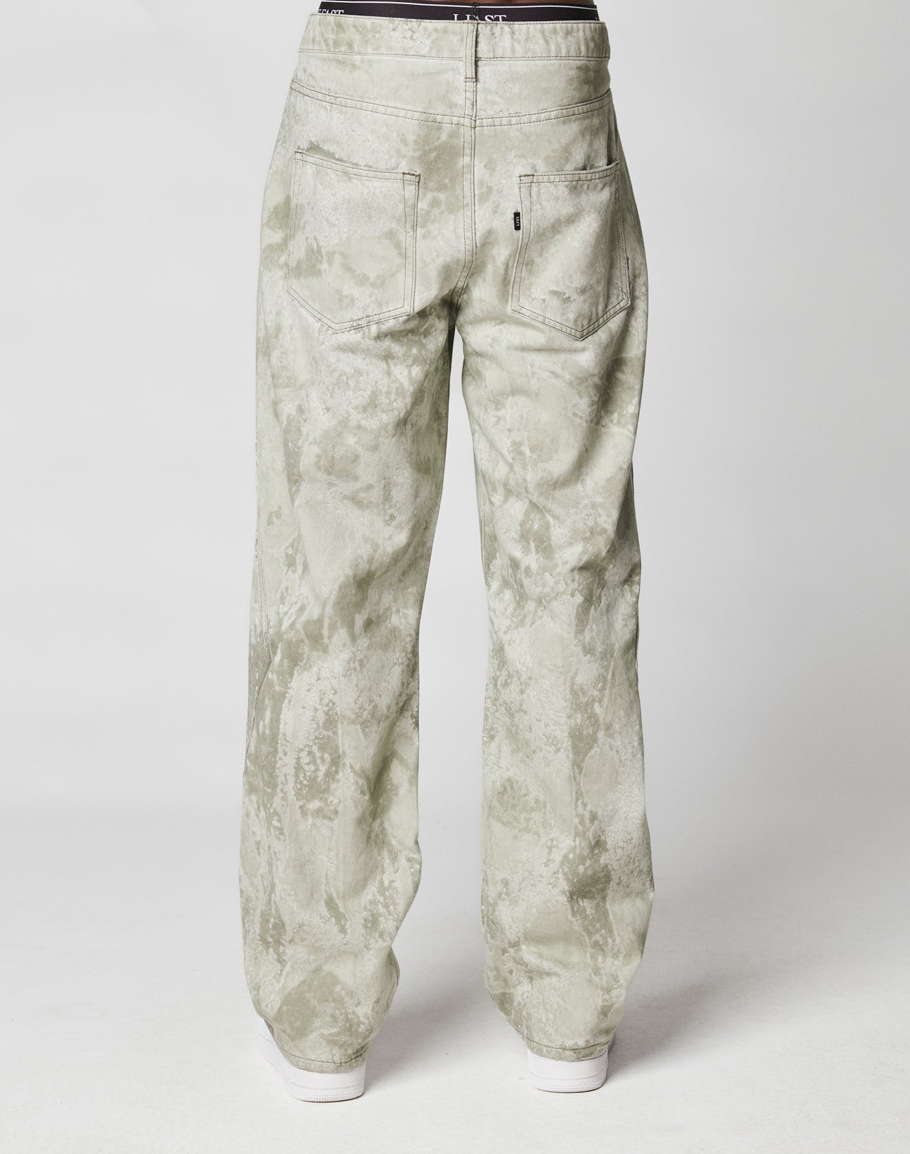 A person is seen from behind wearing LIVE FAST DIE YOUNGs Rhomb Wide Denim—light gray, loose-fitting acid-wash jeans with diamond-shape panels, two back pockets, 100% Baumwolle—draped over white shoes against a plain white background.