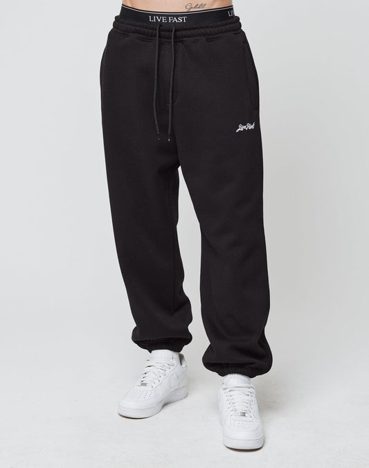 A person wears black Loose Jogger sweatpants by LIVE FAST DIE YOUNG, featuring Live Fast text on the waistband and thigh, elastic cuffs, paired with white sneakers, standing against a plain white background.