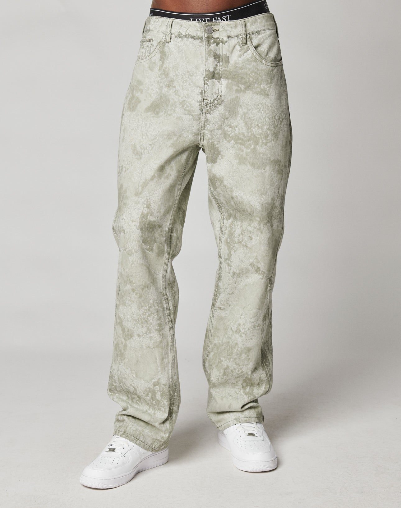 A person wears LIVE FAST DIE YOUNGs Rhomb Wide Denim—light grey/white, marble-washed, baggy jeans with diamond panels and a black “LANDY EAST” waistband—plus white sneakers, standing against a plain light background. Upper body not visible.