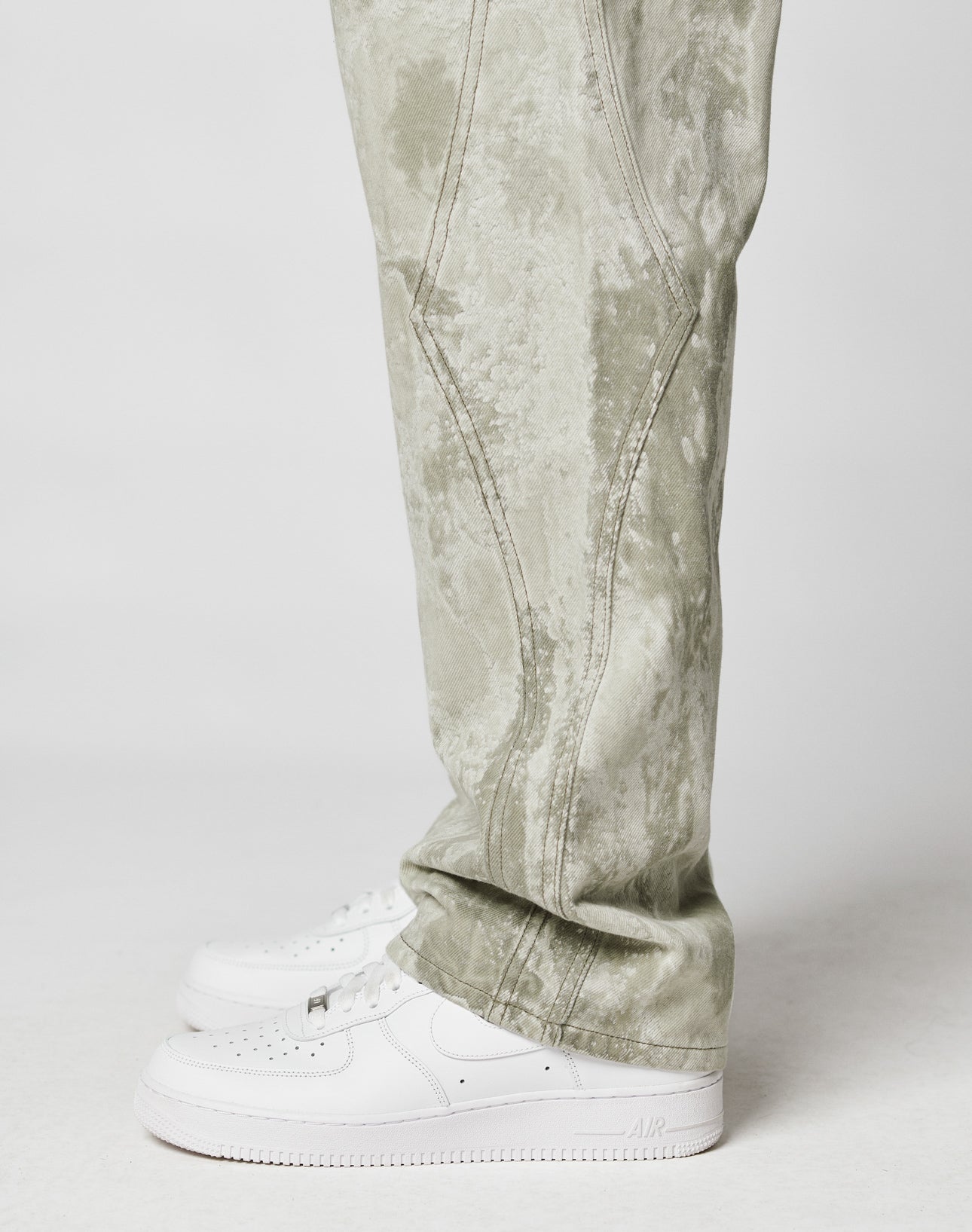A person in LIVE FAST DIE YOUNG Rhomb Wide Denim acid-wash, light green baggy pants and white Nike Air Force 1s, photographed from the side against a plain white background.