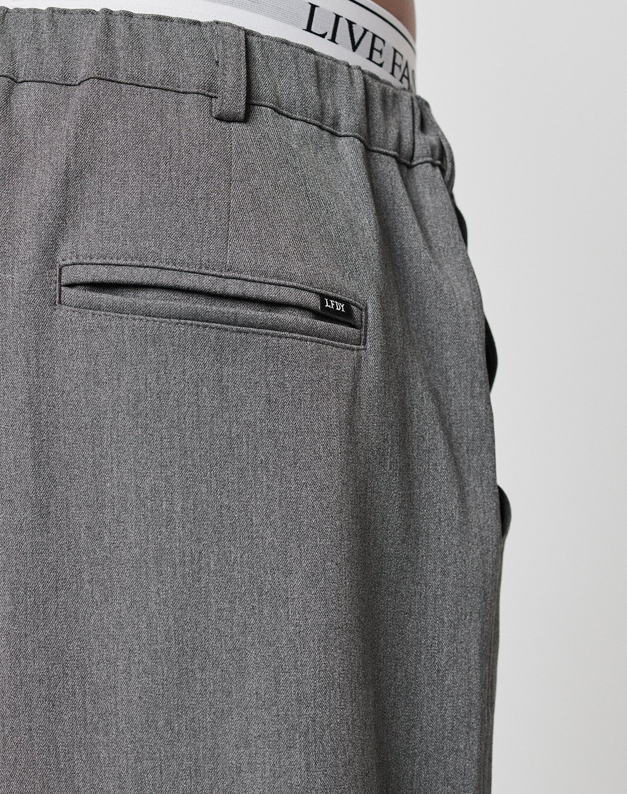 Close-up of the back of gray Wide Casual Pants by LIVE FAST DIE YOUNG with a straight leg and welt pocket. The white underwear’s elastic waistband, featuring LIVE FAST, is visible above the pants.
